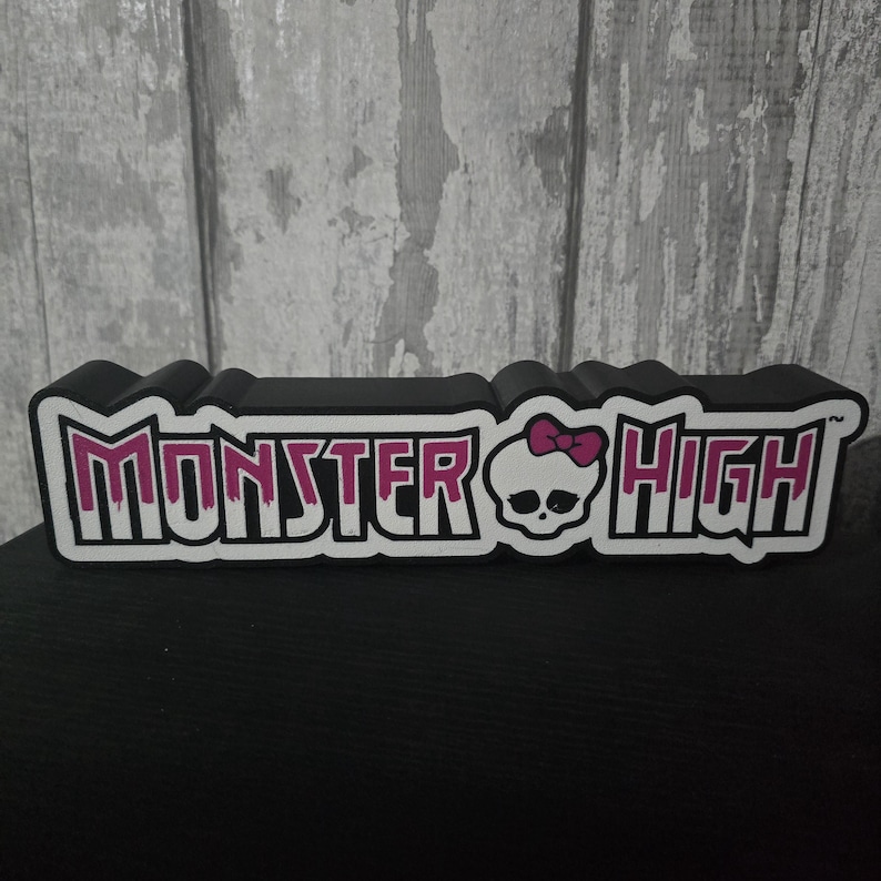 May include: A black, rectangular Monster High logo with white and pink lettering. The logo features a skull with a pink bow. The background is a light gray wooden panel.