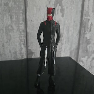 May include: A figurine of a person in a black and red outfit, including a mask and a jumpsuit. The figure is standing with hands at the sides, and the outfit has white accents. The background is a gray and white wall.