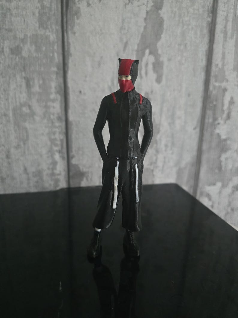 May include: A figurine of a person in a black and red outfit, including a mask and a jumpsuit. The figure is standing with hands at the sides, and the outfit has white accents. The background is a gray and white wall.
