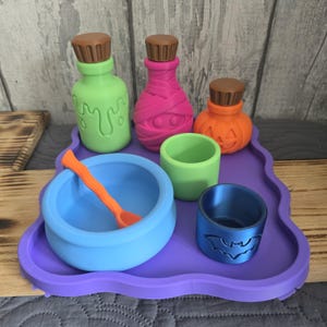 May include: A purple tray holding a collection of small items including a light green bottle with a drip design, a pink mummy-wrapped bottle, an orange pumpkin-shaped bottle, a light blue bowl with an orange spoon, a green cup, and a blue cup with a bat.