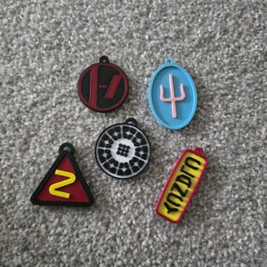 Puede incluir: Cinco llaveros coloridos con varios diseños. Uno es un círculo rojo con una barra negra, otro es un óvalo azul con un tridente rosa, y un tercero es un círculo blanco y negro. Los otros son un triángulo rojo con un "2" amarillo y un rectángulo amarillo con texto negro.