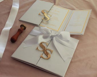 May include: Elegant wedding invitations with gold accents. The invitations are light gray with gold trim and a gold initial monogram. A white ribbon and bow secure the closed invitation. A wooden handled wax stamp and white ribbon are also in the image.