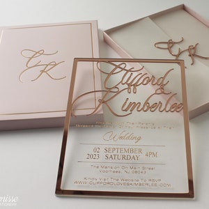 Puede incluir: Una invitaci&oacute;n de boda en oro rosa con los nombres "Clifford & Kimberlee" en escritura elegante. La invitaci&oacute;n incluye la fecha, hora y lugar. Tambi&eacute;n se ve una caja y unas iniciales a juego.