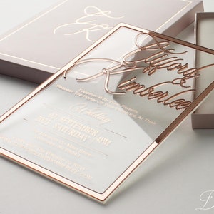 Puede incluir: Una invitaci&oacute;n de boda de acr&iacute;lico transparente con detalles en oro rosa. La invitaci&oacute;n presenta una escritura elegante con los nombres "Lygord & Kimberlee" y detalles de la boda. La invitaci&oacute;n se muestra con una caja a juego.