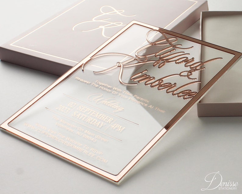 Puede incluir: Una invitaci&oacute;n de boda de acr&iacute;lico transparente con detalles en oro rosa. La invitaci&oacute;n presenta una escritura elegante con los nombres "Lygord & Kimberlee" y detalles de la boda. La invitaci&oacute;n se muestra con una caja a juego.