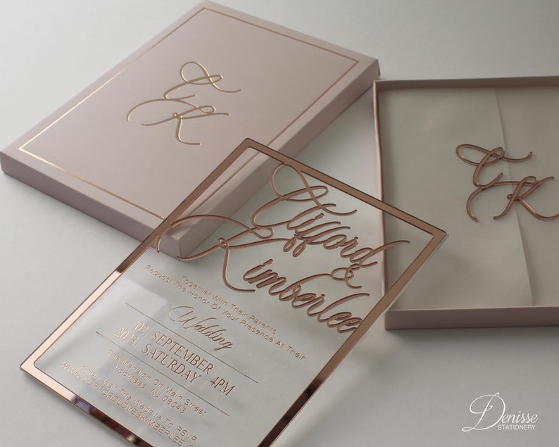 Puede incluir: Una invitaci&oacute;n de boda en oro rosa con una superposici&oacute;n de acr&iacute;lico transparente, con los nombres "Clifford & Kimberlee" en escritura elegante. La invitaci&oacute;n incluye detalles del evento y se presenta en una caja rosa rubor con iniciales a juego.