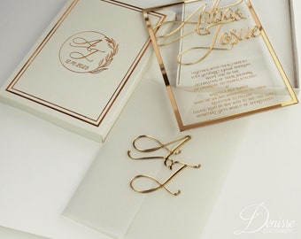 May include: Gold wedding invitation with a clear acrylic cover and a gold border. The invitation features the couple's names, "Athas & Josue", and the date "December 16th, 2025". The invitation also includes the location of the wedding, "Arritola Ranch", and the reception to follow. The invitation is decorated with gold lettering and a gold floral design.