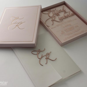Puede incluir: Un juego de invitaciones de boda rosa rubor con detalles dorados. El juego incluye una caja con un monograma dorado, una invitaci&oacute;n cortada con l&aacute;ser y un sobre de vitela. La invitaci&oacute;n presenta los nombres "Tiffany & Kimberlee" y detalles de la boda.