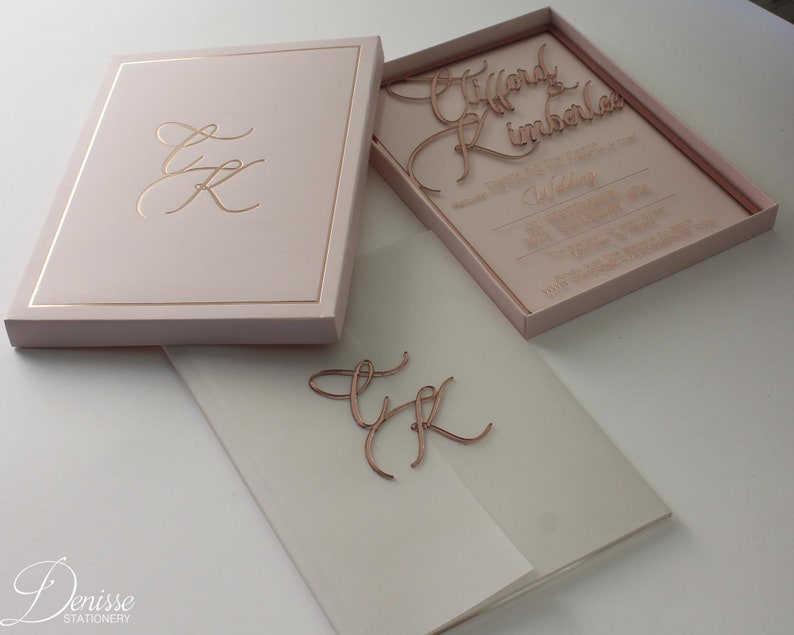 Puede incluir: Un juego de invitaciones de boda rosa rubor con detalles dorados. El juego incluye una caja con un monograma dorado, una invitaci&oacute;n cortada con l&aacute;ser y un sobre de vitela. La invitaci&oacute;n presenta los nombres "Tiffany & Kimberlee" y detalles de la boda.