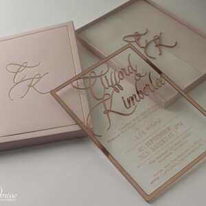 Puede incluir: Un conjunto de invitaciones de boda rosa rubor con una invitaci&oacute;n de acr&iacute;lico transparente y letras en oro rosa. La invitaci&oacute;n incluye los nombres "Clifford & Kimberlee", detalles de la boda y un sitio web. Tambi&eacute;n se incluye una caja a juego con iniciales doradas.