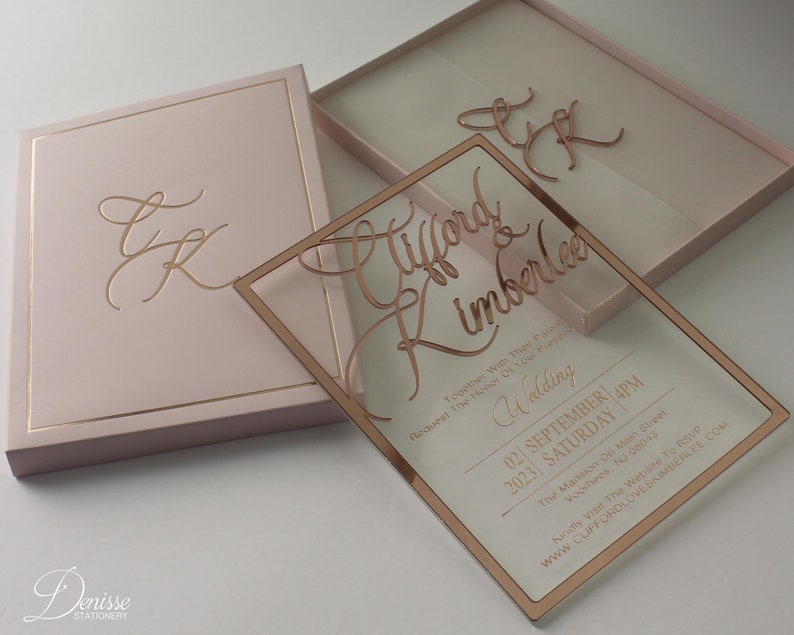 Puede incluir: Un conjunto de invitaciones de boda rosa rubor con una invitaci&oacute;n de acr&iacute;lico transparente y letras en oro rosa. La invitaci&oacute;n incluye los nombres "Clifford & Kimberlee", detalles de la boda y un sitio web. Tambi&eacute;n se incluye una caja a juego con iniciales doradas.