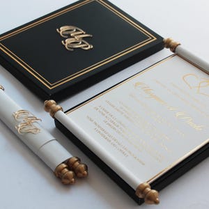 Könnte beinhalten: Eine weiße Schriftrolle mit goldenen Verzierungen und eine schwarze Box mit goldenen Verzierungen. Die Schriftrolle trägt eine goldene Aufschrift "Maspored Paula" und die Box eine goldene Aufschrift "C&P".