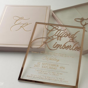 Puede incluir: Elegante juego de invitaciones de boda con una invitaci&oacute;n de acr&iacute;lico transparente con letras doradas y una caja a juego. La invitaci&oacute;n incluye los nombres "Clifford & Kimberlee", la fecha, la hora y los detalles del lugar. La caja es rosa rubor con detalles dorados.