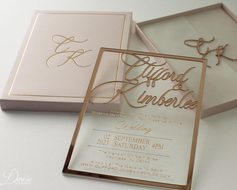Puede incluir: Elegante juego de invitaciones de boda con una invitaci&oacute;n de acr&iacute;lico transparente con letras doradas y una caja a juego. La invitaci&oacute;n incluye los nombres "Clifford & Kimberlee", la fecha, la hora y los detalles del lugar. La caja es rosa rubor con detalles dorados.