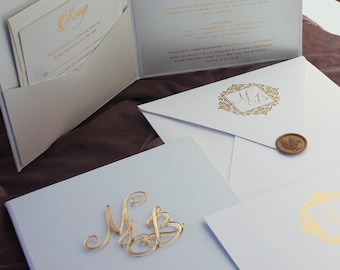May include: White wedding invitation suite with gold foil accents. The RSVP card is in a pocket on the front of the invitation. The envelope has a wax seal with a floral design. The invitation features the initials "MB" in a gold frame.