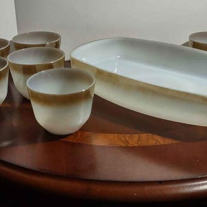 May include: A set of ceramic serving dishes and cups. The set includes a large, oval-shaped serving dish and several small cups. The dishes and cups are white with a brown band around the rim. The set is arranged on a dark wooden surface.