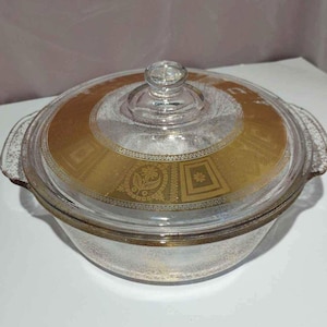 May include: A clear glass casserole dish with a matching lid. The lid and the rim of the dish are decorated with a gold floral and geometric pattern. The dish has two handles and a clear knob on the lid.