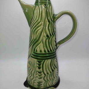 May include: A tall, green ceramic pitcher with a unique, textured design. The pitcher features a spout, a handle, and a decorative pattern of curved lines. The base is dark brown, and the overall aesthetic is artistic and handcrafted.