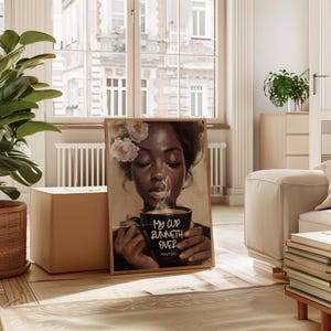 May include: A framed art print featuring a portrait of a person with flowers in their hair, holding a black mug with the text "My cup runneth over" and the Bible verse Psalm 23:5. The artwork has a warm color palette and is displayed in a well-lit room.