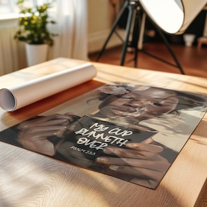 May include: A print featuring a woman holding a cup with the text "My cup runneth over" and "Psalm 23:5". The artwork has a warm color palette and is displayed on a wooden surface. A rolled-up poster is in the background.