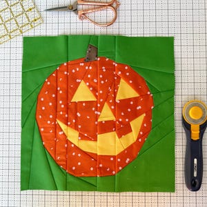 May include: A green fabric square with a patchwork pumpkin in the centre. The pumpkin is orange with white polka dots and a yellow smiling face.