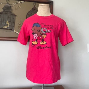 May include: Pink t-shirt with a cartoon image of Mickey Mouse holding a magnifying glass. The text "The Mouse Club" and "Get Disneyized" are printed on the shirt.