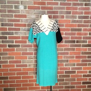 May include: A turquoise dress with a black and white geometric pattern on the neckline and short sleeves. The dress is on a mannequin against a brick wall.