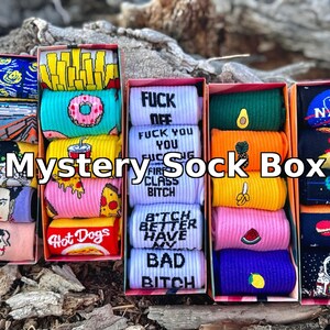 Può includere: Quattro scatole colorate di calzini con diversi design. Le scatole sono etichettate "Mystery Sock Box". I calzini nelle scatole hanno design come hot dog, pizza, avocado, ananas e un logo NASA.