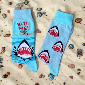 以下が含まれることがあります： 青い靴下2足。片方の靴下にはサメのグラフィックと「Bite Me!」の文字が、もう片方の靴下にはサメのグラフィックが描かれています。