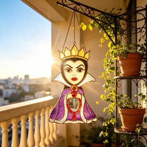 Puede incluir: Atrapasoles de la Reina Malvada de vidrieras con corona dorada, capa morada y detalles rojos. El atrapasoles cuelga de una cadena y está suspendido de un soporte de metal negro. El fondo muestra un balcón y un paisaje urbano.