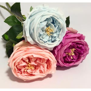 May include: A bouquet of artificial roses in shades of light blue, peach, and magenta. The roses have multiple layers of petals and yellow centers. Green leaves and stems are visible.