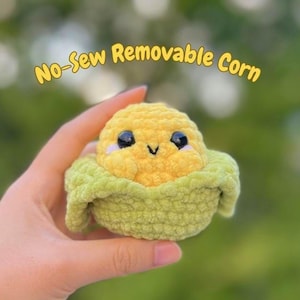 Könnte beinhalten: Ein handgefertigtes, gehäkeltes Mais-Plüschtier mit einem lächelnden Gesicht, gehalten in einer Hand. Der Mais ist gelb mit schwarzen Augen und einem weißen Highlight, eingebettet in eine grüne Hülle. Der Text oben lautet "No-Sew Removable Corn".