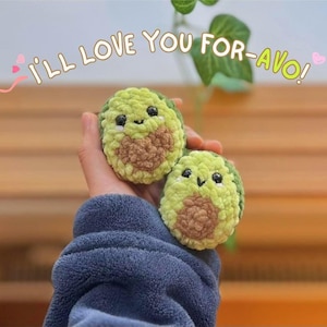 May include: Two handmade crochet avocado plushies with smiling faces. The avocados are green and brown, with black eyes and a smiling mouth. The text "I'll love you for-avo!" is written above the avocados.