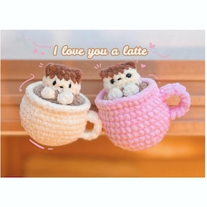 May include: Two crocheted coffee mugs with cute animal faces peeking out of the top. The mugs are in the colours pink and cream and the text "I love you a latte" is written above them.