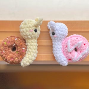 Könnte beinhalten: Zwei gehäkelte Schnecken-Plüschtiere mit Donut-Schalen. Eine Schnecke ist weiß mit einer rosafarbenen Donut-Schale mit weißen Streuseln. Die andere Schnecke ist gelb mit einer braunen Donut-Schale mit bunten Streuseln.