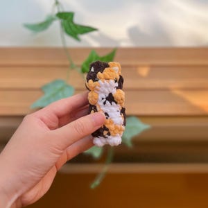 May include: A handmade crochet cat toy in a cylindrical shape. The toy features a pattern of brown, white, and yellow, resembling a calico cat. The crochet stitches are visible, giving it a textured appearance. The toy is held in a hand.