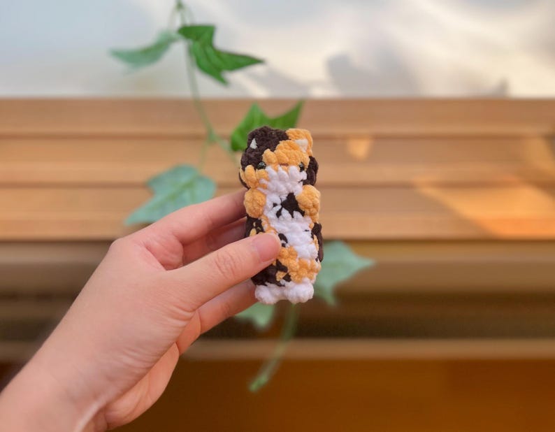 May include: A handmade crochet cat toy in a cylindrical shape. The toy features a pattern of brown, white, and yellow, resembling a calico cat. The crochet stitches are visible, giving it a textured appearance. The toy is held in a hand.