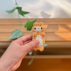 May include: A small, handmade crochet tiger toy in shades of orange and white. The plush toy has black eyes and is held in a person's hand. The background includes green leaves and a wooden surface.