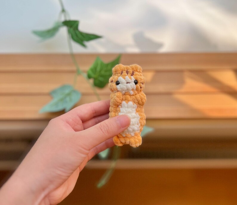 May include: A small, handmade crochet tiger toy in shades of orange and white. The plush toy has black eyes and is held in a person's hand. The background includes green leaves and a wooden surface.