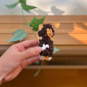 May include: A small, handmade crochet item in shades of brown, tan, white, and black. The item is cylindrical with a textured surface and appears to be a decorative or novelty item. The background includes wooden slats and green leaves.