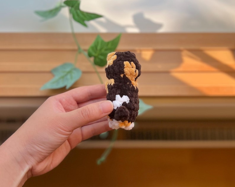 May include: A small, handmade crochet item in shades of brown, tan, white, and black. The item is cylindrical with a textured surface and appears to be a decorative or novelty item. The background includes wooden slats and green leaves.
