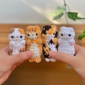 May include: Four handmade crochet cat figurines in various colors: white with brown spots, orange tabby, calico, and white with pink spots. Each cat has black button eyes and a simple, rounded shape. The cats are held in front of a blurred background.