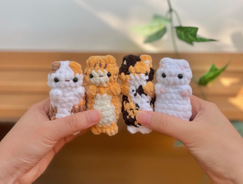 May include: Four handmade crochet cat figurines in various colors: white with brown spots, orange tabby, calico, and white with pink spots. Each cat has black button eyes and a simple, rounded shape. The cats are held in front of a blurred background.