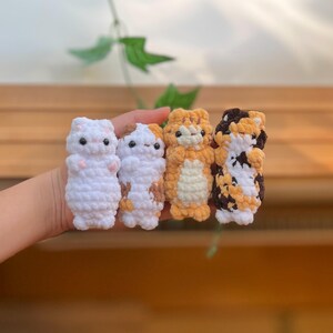 May include: Four handmade crochet cat figurines in white, calico, orange, and calico patterns. Each cat has small black eyes and a cute, upright pose. The yarn is soft and textured, creating a charming, whimsical aesthetic.