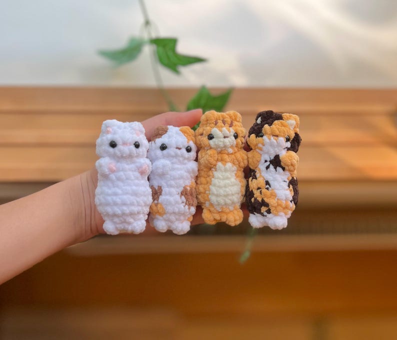 May include: Four handmade crochet cat figurines in white, calico, orange, and calico patterns. Each cat has small black eyes and a cute, upright pose. The yarn is soft and textured, creating a charming, whimsical aesthetic.