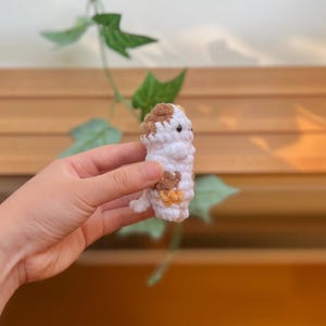 May include: A handmade crochet animal in white, brown, and yellow. The small, stuffed creature has a rounded body, brown spots, and small black eyes. It is held in a person's hand against a blurred background of wood and greenery.