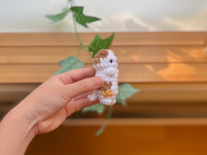 May include: A handmade crochet animal in white, brown, and yellow. The small, stuffed creature has a rounded body, brown spots, and small black eyes. It is held in a person's hand against a blurred background of wood and greenery.