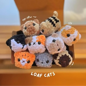 May include: A selection of handmade, crocheted cat-shaped toys in a variety of colours, including orange, black, white, and grey. The toys are arranged in a wooden box. The text "LOAF CATS" is visible at the bottom.