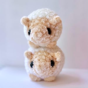 May include: Two plush white and cream colored sheep toys stacked on top of each other. The top sheep has a white face and the bottom sheep has a cream colored face. Both sheep have black eyes.