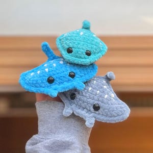May include: Three crocheted plush toys shaped like stingrays. The top stingray is teal blue with white spots. The middle stingray is light blue with white spots. The bottom stingray is gray with white spots.